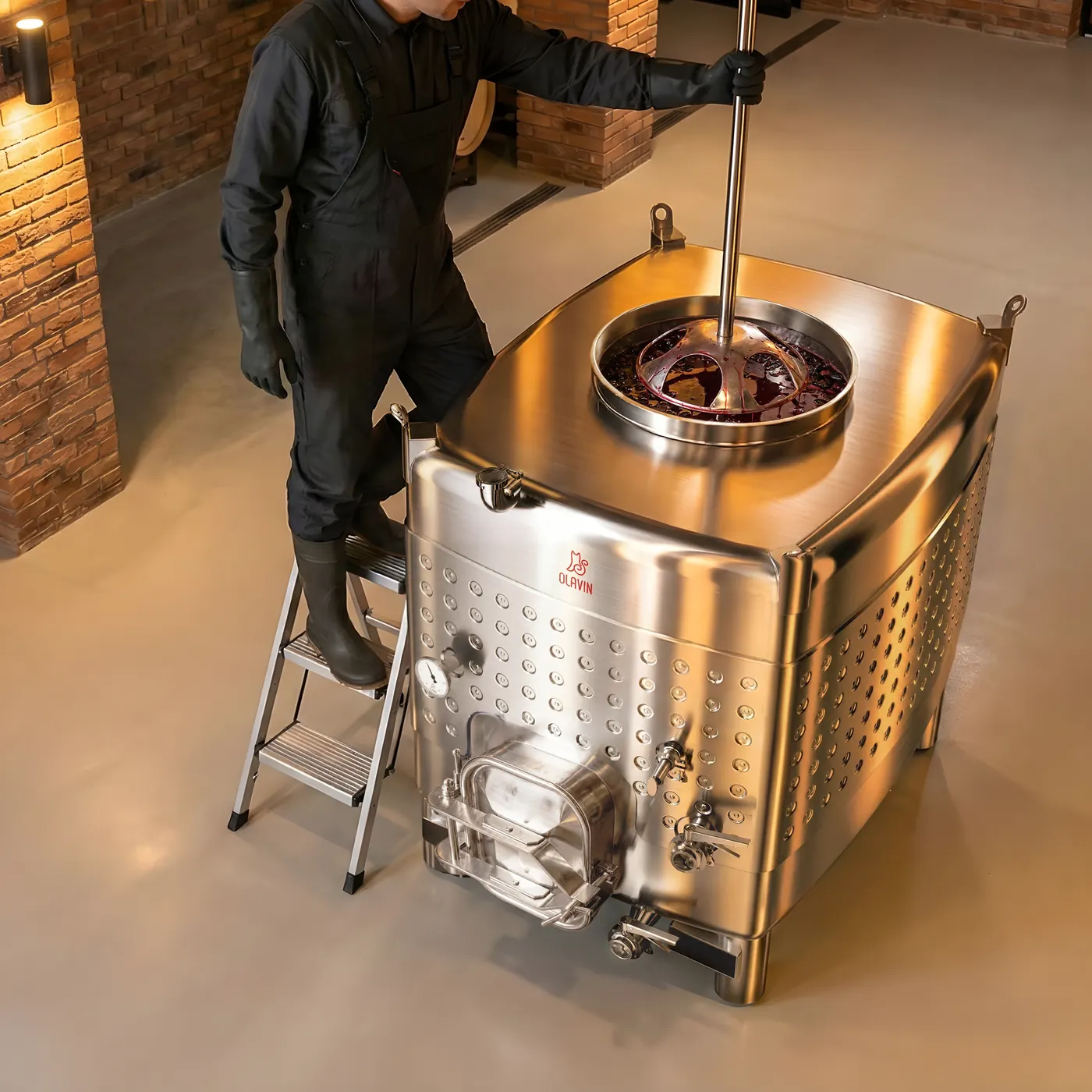 A winemaker on a step ladder punching down through the top entry of an Olavin Mini Tanker, the cap of skins floating in the open manway
