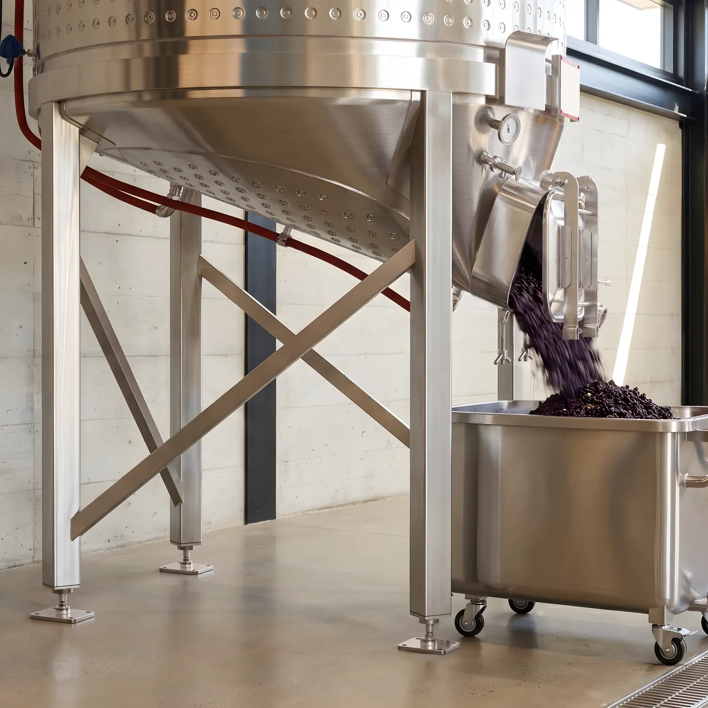 Pomace sliding out of the open hatch at the base of a conical fermenter into a stainless bin on castors, moved by gravity rather than by hand
