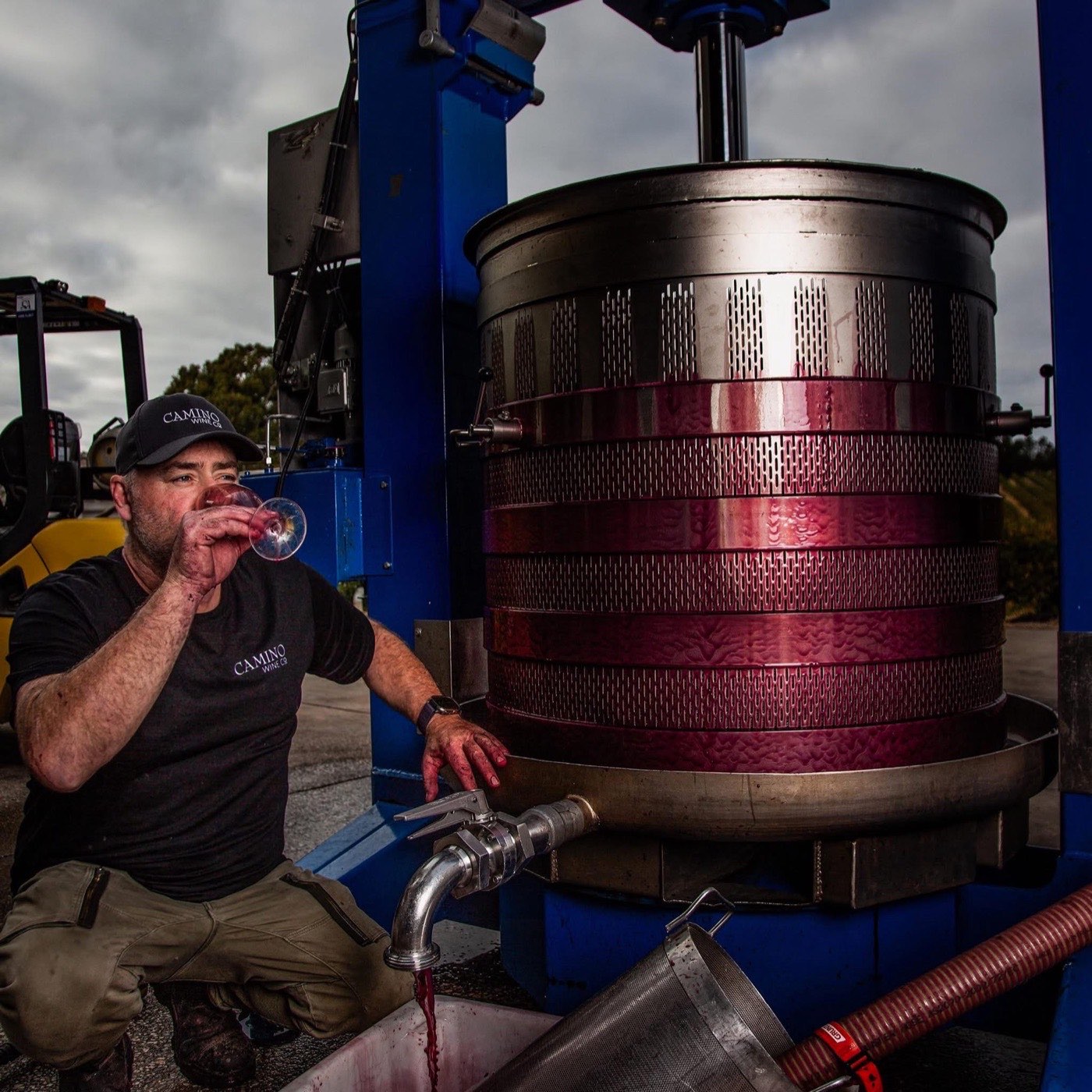 Jason Barrette crouches beside a loaded basket press at Camino Wine Co, tasting a glass of young Shiraz as pressed wine runs from the outlet into a bucket.