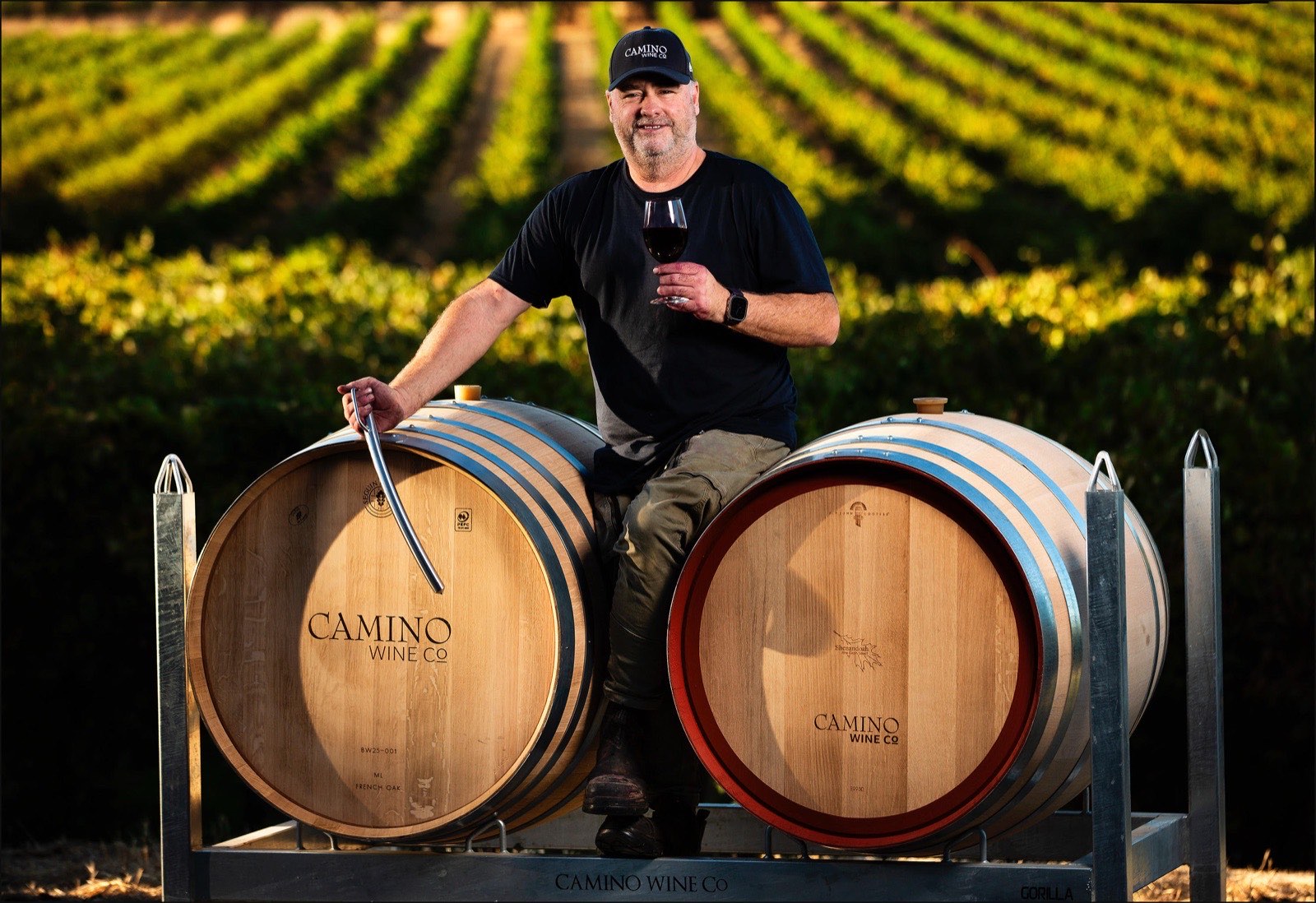 Jason Barrette sits on a pair of Camino Wine Co French oak barrels in front of Barossa Valley vineyard rows, holding a glass of red wine and a wine thief.
