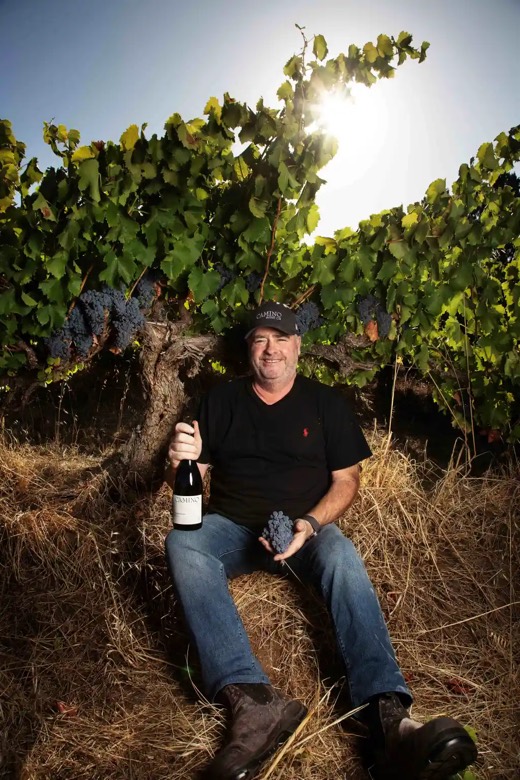 Jason Barrette, winemaker and co-founder of Camino Wine Co, seated against an old Barossa Valley vine holding a bottle and a bunch of Shiraz.