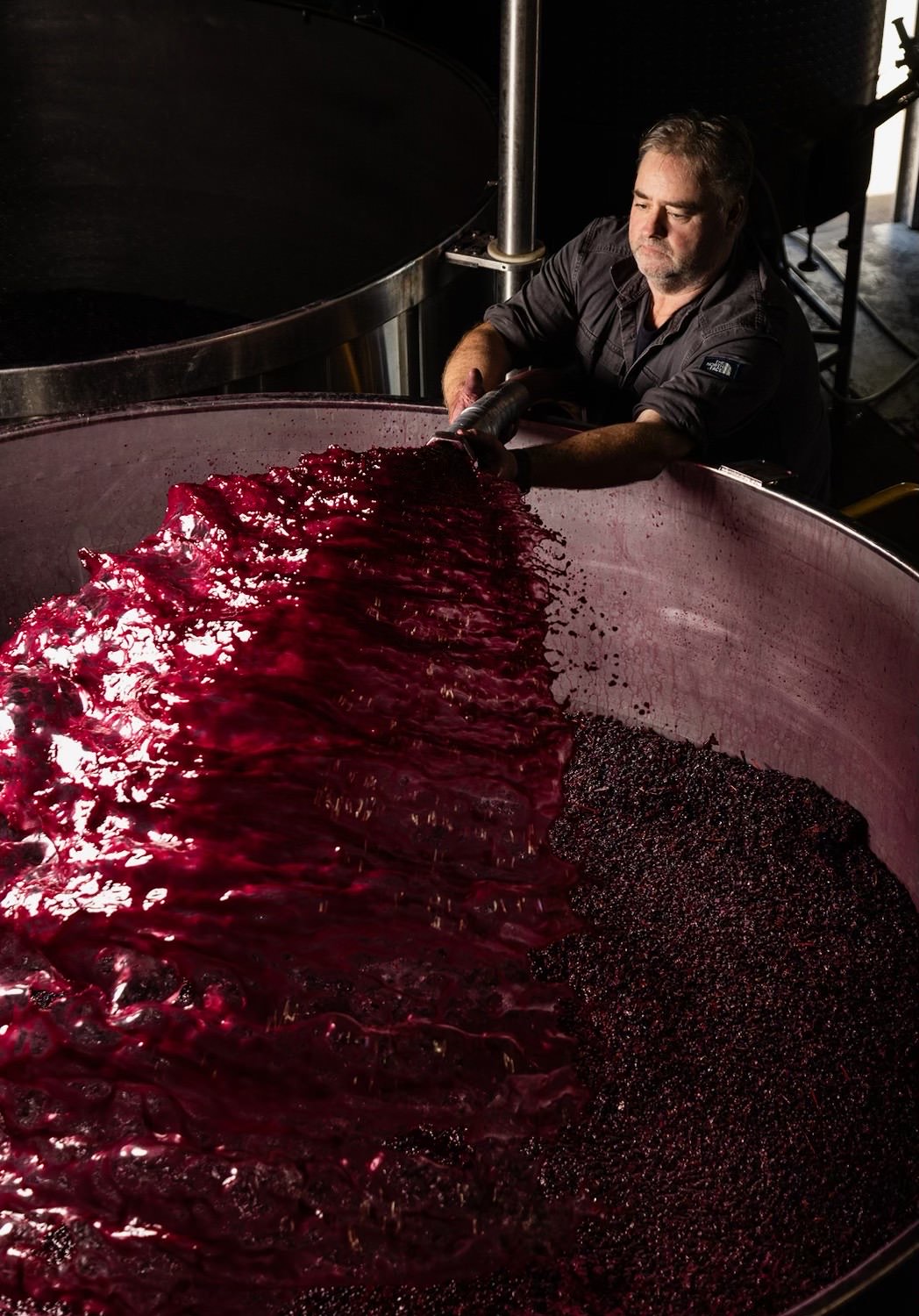 Jason Barrette leans over the open top of an Olavin conical fermenter, using a manual heading-down board to submerge a Shiraz cap into the bright red fermenting must.